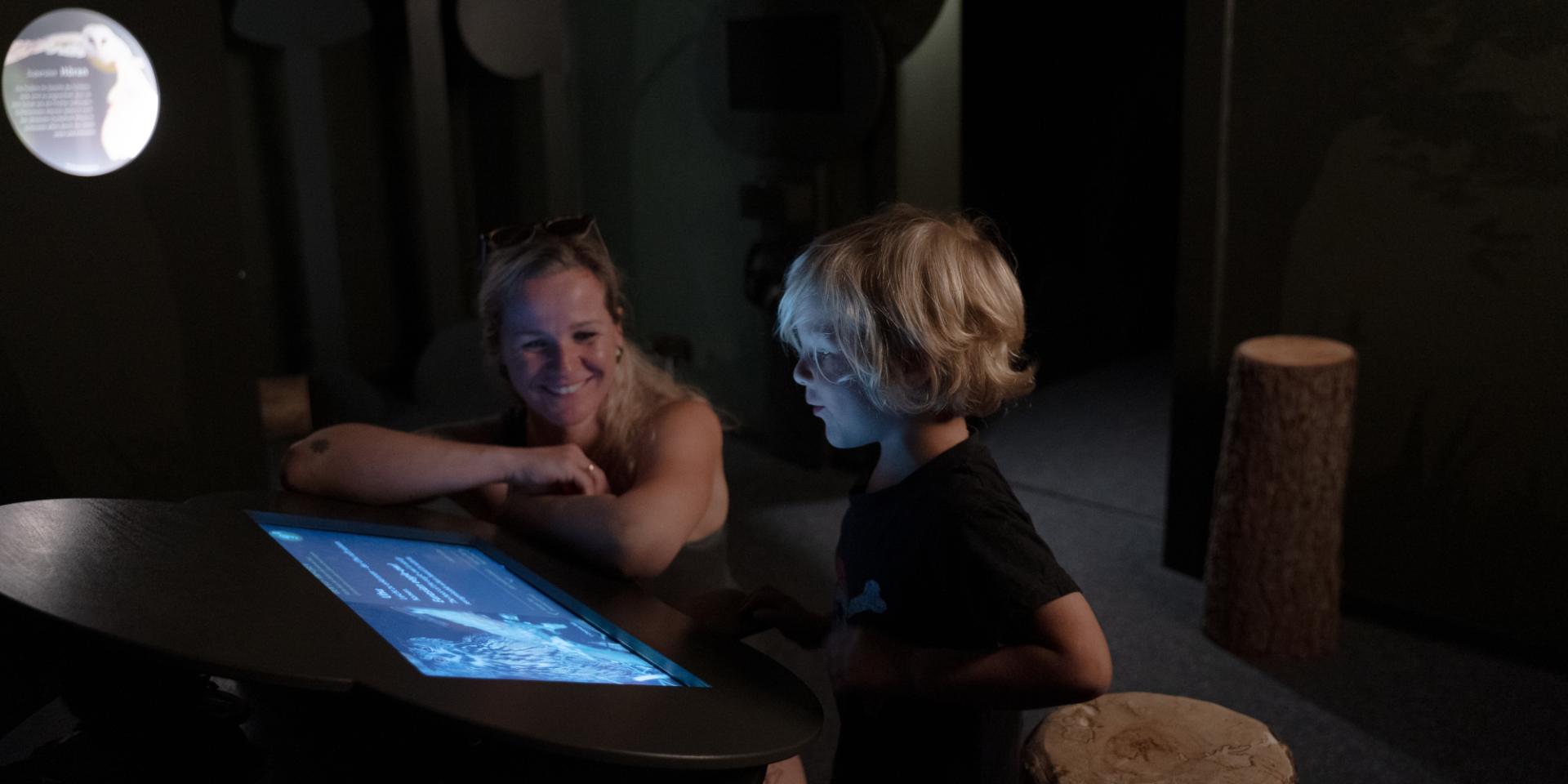 Eine junge Frau lacht in einem dunklen Raum ihren Sohn an, der auf einen Bildschirm schaut, im Hintergrund ist ein Vollmond zu sehen
